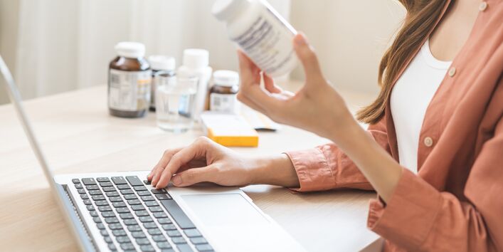 Photo of a young woman using a laptop, medication in one hand, looking up information online, with more medication on a table in the background 