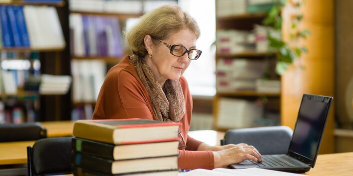 Eine Frau sitzt mit einem Laptop in einer Bibliothek und liest ein Buch.