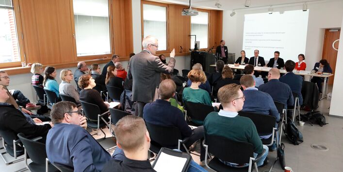 Dies ist ein Bild von der Fachtagung 2019: Mit wissenschaftlichen Methoden die Klimaanpassung unterstützen, welches im Ausstellungsraum im Umweltbundesamt aufgenommen worden ist. Das Bild zeigt Personen die auf gestühlten Reihen sitzen und ndach vorne auf eine Präsentation schauen - ein klassischer Vortrag. Ein älterer Mann steht allerdings und stellt eine Frage an die Referenten. Im Hintergrund ist eine PowerPoint zu erkennen.