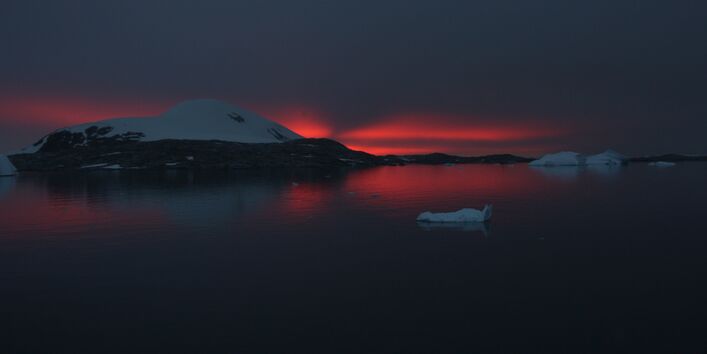Es ist schon fast Nacht über der Antarktis. Nur über dem Horizont ist noch ein rotes Licht zu sehen. Es spiegelt sich im Wasser. So sieht man noch Eisschollen und einen schneebedeckten Hügel.