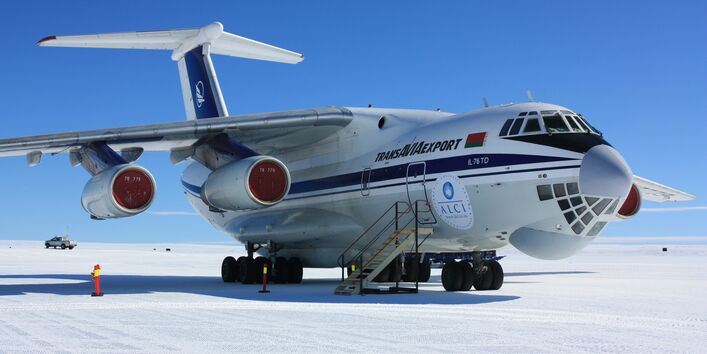 Ein weißes Transportflugzeug mit blauen Streifen an der Seite steht auf der Start- und Landebahn in der Antarktis. Eine Treppe steht vor der Tür. Im Hintergrund fährt ein Pick-up.
