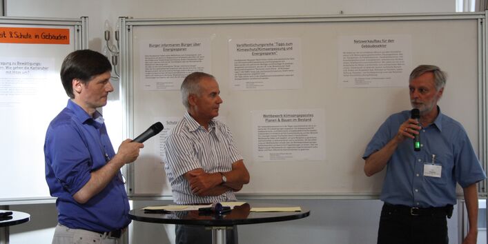 Vor einer weißen Tafel mit aufgeklebten Blättern mit Texten stehen drei Männer. Zwei der drei Männer halten ein Mikrofon. Der eine spricht in dieses hinein. In der Mitte ist ein Stehtisch.