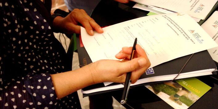 Es ist ein Stehtisch zu sehen. Eine Person steht an diesem, dabei ist der Oberkörper bis zum Hals zu sehen. Die Person hält einen Stift in der Hand und füllt einen Anmeldebogen aus. Daneben tut es ihr eine Person gleich, von der nur eine Hand, Stift und Zettel zu sehen sind.