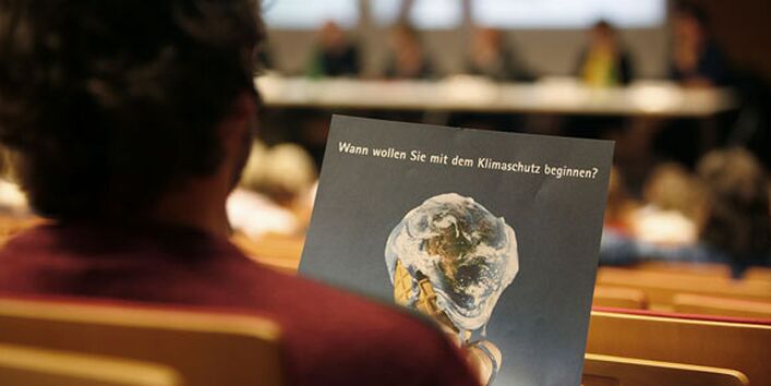 Ein Zuhörer blickt auf eine Broschüre. Das Titelbild zeigt eine Eistüte mit einem verlaufenden Erdball als Weltkugel, darüber steht: "Wann wollen Sie mit dem Klimaschutz beginnen?"