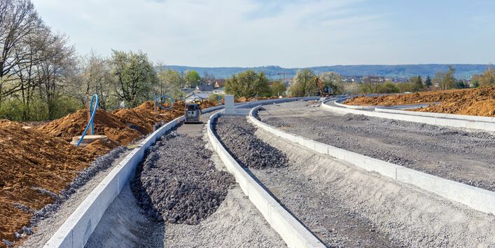 Das Bild zeigt einen Straßenneubau. Z sehen ist das teil-fertige Fundament für die Straße. Im Vordergrund ist es noch in Arbeit, im Hintergrund schon verdichtet. Links von der neuen Straße sind Erdhaufen zu sehen, dahinter eine Baumreihe. Im Hintergrund sind die Dächer eines Wohngebiets zu erkennen.