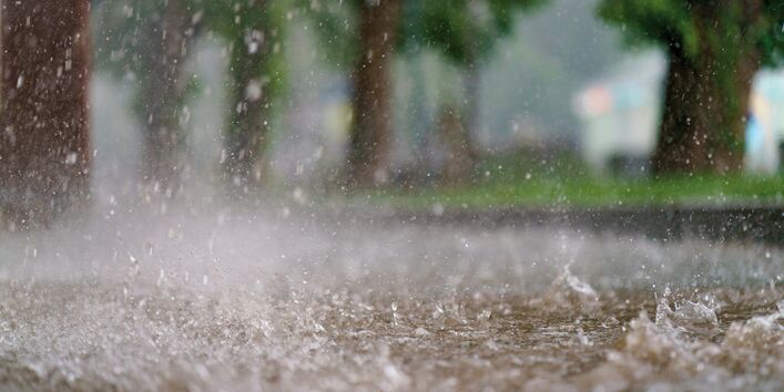 The picture shows a path or a road at the time of a heavy rain event. There is standing water visible on the path. The falling raindrops cause the water to splash up from the ground. In the background, it is just possible to make out a blurred line of trees.