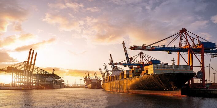 The picture shows a large container port beneath a low sun. Visible in the foreground are a laden container ship and a few loading cranes. In the background there is another container ship docked at the port.
