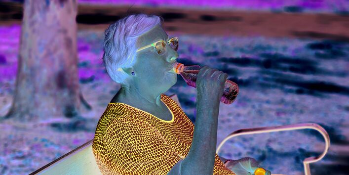 Das Bild zeigt eine ältere Frau, die auf einer Parkbank im Schatten unter einem Baum sitzt und aus einer Wasserflasche trinkt.