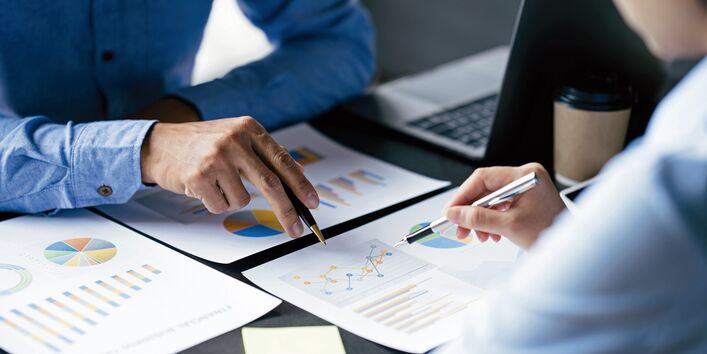 Das Bild zeigt die Arme und Hände zweier förmlich gekleideter Personen, die an einem Schreibtisch sitzen. Auf dem Tisch befinden sich Dokumente, die verschiedene Diagramme zeigen, Klebezettel sowie ein Laptop. Beide Personen halten in je einer Hand einen Kugelschreiber, mit dem sie auf dasselbe Liniendiagramm deuten.