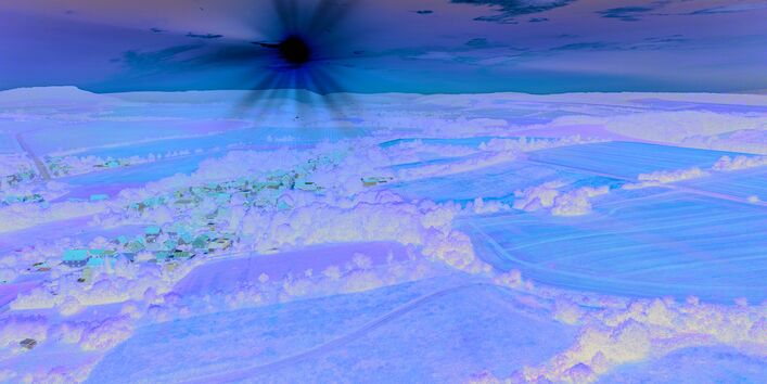 Das Bild zeigt eine leicht hüglige Landschaft, die ackerbaulich geprägt ist. In der Bildmitte ist ein Dorf in die Landschaft eingebettet. Am fast wolkenlosen, zum Teil schon orange gefärbten Himmel steht eine tiefstehende Sonne, die die Landschaft in warmes Licht hüllt.