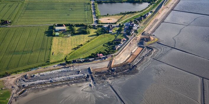 The picture shows an aerial view of a land protection dyke under construction. In the background, there are agricultural areas and houses as well as standing water visible. 