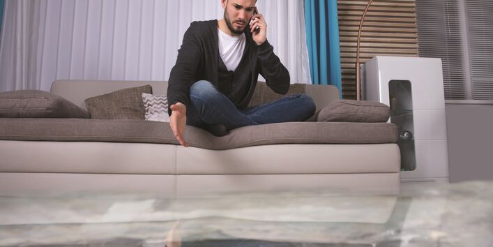 The picture shows a man sitting cross-legged on a sofa and talking on the phone. There is water on the floor of the room. The man points reproachfully at the surface of the water.