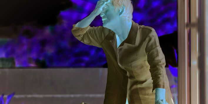 Das Bild zeigt eine ältere Frau mit weißer Bluse. Die Dame hält sich mit einer Hand an ihrem Rollator fest, den Handrücken der anderen Hand hält sie an ihre Stirn. Im Hintergrund sind verschwommen Baumkronen oder Sträucher in hellem Licht zu sehen.