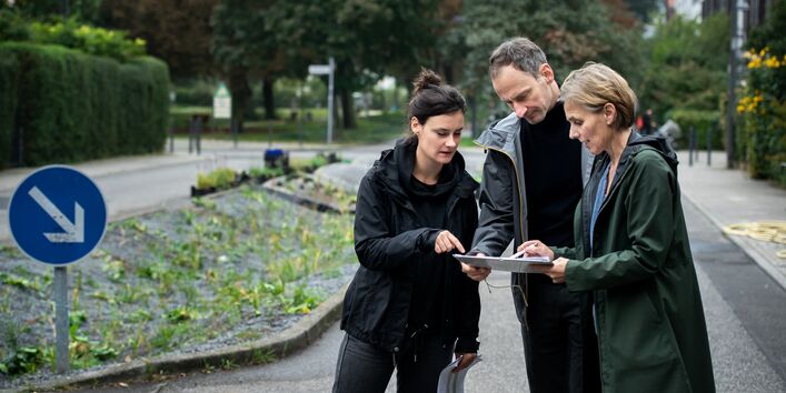 Der Wettbewerb Blauer Kompass stellt seine Bewerber vor, damit abgestimmt werden kann, welches Projekt gewinnt. Zu sehen sind drei Menschen der Berliner Regenwasseragentur, die auf der Straße stehen uns sich einen Plan angucken. 