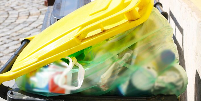 Zu sehen ist die gelbe Tonne, in die der Plastikabfall kommt. Die Mülltonne ist überfüllt. Aus diesem Grund ist die Tonne nicht richtig verschlossen und ein Müllbeutel mit Plastikabfall ist zu sehen. 
