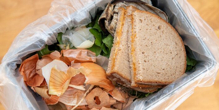 Ein Mülleimer mit Lebensmittelresten. Zu sehen sind Zwiebelschalen und zwei geschmierte Brote. 