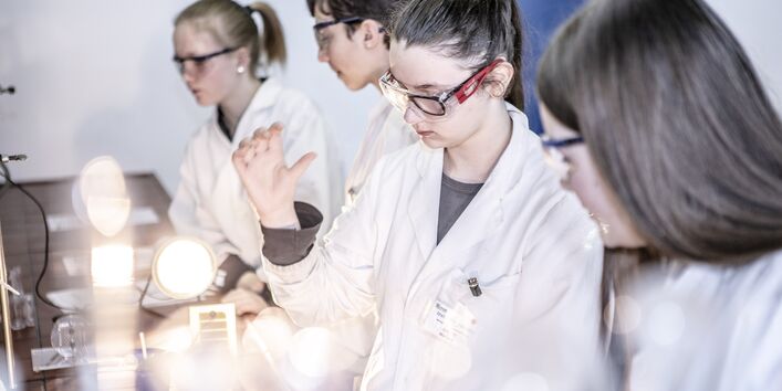 Jugendliche Mädchen und Jungen in einem Labor mit Kitteln und Schutzbrillen beim Zukunftstag in einem Labor. 