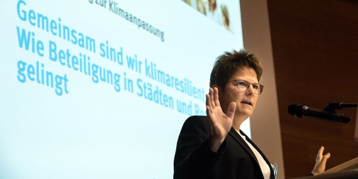 Teilnehmerin steht am Rednerpult, trägt vor und gestikuliert. Im Hintergrund ist der Titel der Veranstaltung zu sehen: Gemeinsam sind wir klimaresilient - Wie Beteiligung in Städten und Regionen gelingt