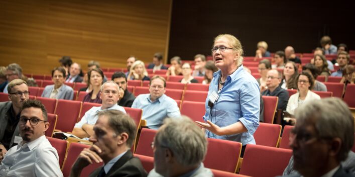 Teilnehmerin steht präsent im Hörsaal innerhalb der roten Stuhlreihen und spricht. Die anderen Teilnehmerinnen und Teilnehmer sitzen rundherum und hören zu.