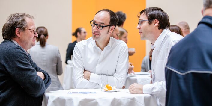 Teilnehmende stehen an weißen Stehtischen in der Pause unterhaltend. Zwei Teilnehmde tragen weiße Hemden. Der helle Raum ist mit gelber Farbe abgesetzt.