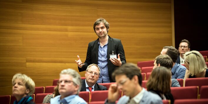  Teilnehmer steht präsent im Hörsaal innerhalb der roten Stuhlreihen und spricht. Die anderen Teilnehmerinnen und Teilnehmer sitzen rundherum und hören zu.