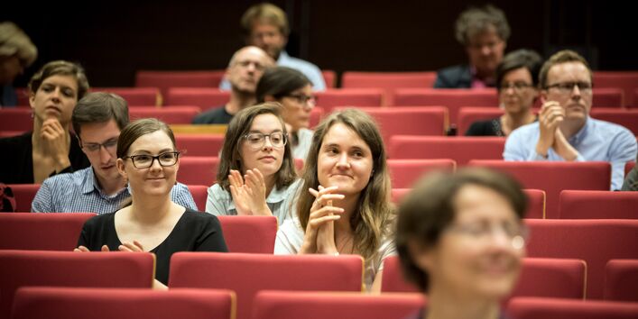Teilnehmerinnen und Teilnehmer sitzen applaudierend in den roten Stuhlreihen des Hörsaales.
