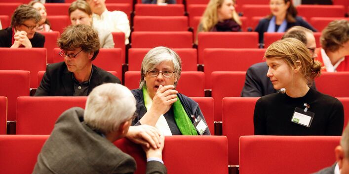 Im Hörsaal sitzen die Teilnehmenden auf den roten Sitzen und diskutieren miteinander. Eine Teilnehmende mit grünem Tuch um dem Hals gestikuliert dabei mit der rechten Hand.