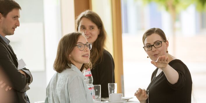 Teilnehmer und Teilnehmerinnen stehen in der Pause am Bistrotisch. Sie folgen schauend dem Hinweis einer Teilnehmerin, die in eine Richtung zeigt. Auf dem Bistrotisch stehen Gläser, Tassen und Teller sowie ein Getränkeflasche.Auf dem B