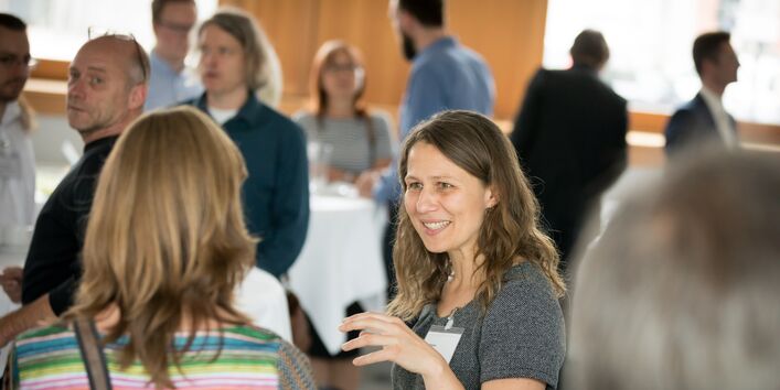 Zwei Teilnehmerinnen unterhalten sich gestikulierend. Im Hintergrund stehen weitere Teilnehmerinnen und Teilnehmer während der Pause ebenfalls an den Bistrotischen.