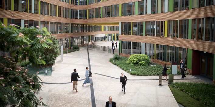 Blick von einer Brücke im Atrium des Umweltbundesamtes auf Teilnehmerinnen und Teilnehmer auf dem Weg zu den Workshopräumen. Das Atrium ist begrünt, die Innenfassade besteht aus Holz und Glas.