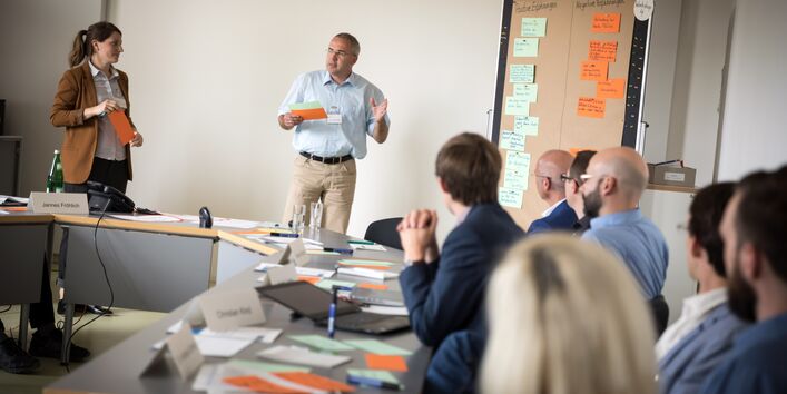 Vortragender steht an mit Moderationskarten bestückte Moderationswand und hält beschriftete orangene und grüne Moderationskarte in der Hand und spricht in den Raum, wo andere Teilnehmerinnen und Teilnehmer am Tisch in Reihe sitzend zuhören. Im Hintergrund steht eine weitere Teilnehmerin.