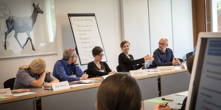 Teilnehmerinnen und Teilnehmer sitzen sich gegenüber in Reihe und diskutieren miteinander. Im Hintergrund steht ein Flipchart. An der Wand hängt ein Bild mit einem Reh darauf.