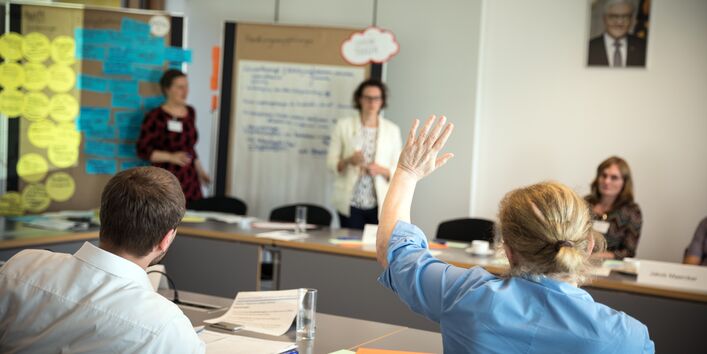 Zwei Teilnehmerinnen stehen vor zwei Moderationswänden. Die Wände sind mit Moderationsmaterialien bestückt. Im Vordergrund hebt eine Teilnehmerin in blauer Bluse die Hand zur Wortmeldung. Andere Teilnehmerinnen und Teilnehmer sitzen sich geben über am Arbeitstisch. Im Hintergrund hängt ein Bild des deutschen Bundespräsidenten.