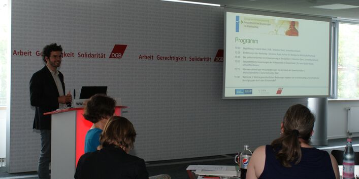 Vortragender steht am Rederpult und präsentiert im Hintergrund das Programm des Stakeholderdialoges.