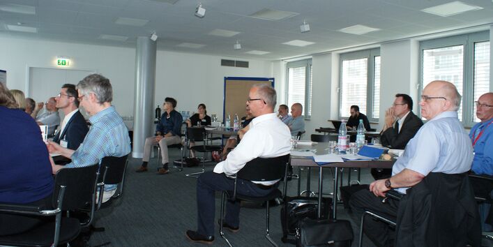 Teilnehmer sitzen im Arbeitsgruppenraum an Arbeitsgruppentischen mit dem Redner zugewandten Gesichtern.