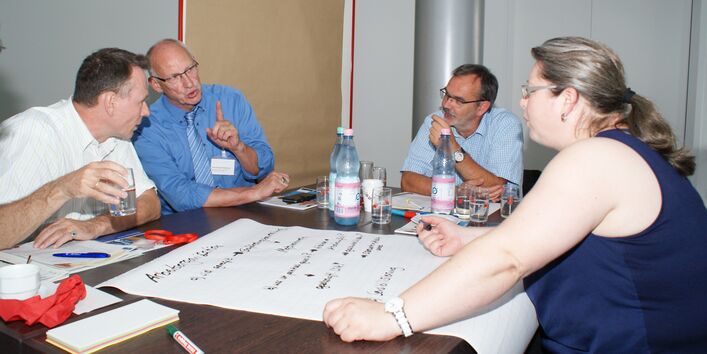 Teilnehmer diskutieren am Gruppenarbeitstisch. Eine Teilnehmerin schreibt Diskussionsergebnisse auf Posterpapier, welches auf dem Tisch liegt. Erfrischungsgetränke stehen auf dem Tisch. Ein Teilnehmer trinkt aus einem Glas Mineralwasser.