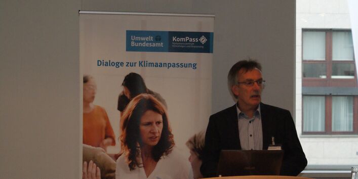 Vortragender steht am Stehpult vor einem aufgeklappten Laptop. Im Hintergrund steht das Rollup Dialoge zur Klimaanpassung. Der Blick geht im Hintergrund rechts durchein Fenster hinaus auf eine Häuserfassade.