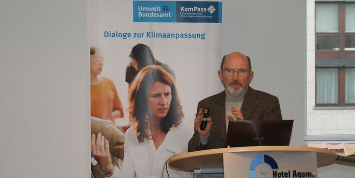 Vortragender steht am Rednerpult vor einem aufgeklappten Laptop und trägt gestikulierend vor. Vorne auf dem Stehpult steht der Name des Tagungszentrums Aquino und im Hintergrund steht das Rollup Dialoge der Klimaanpassung und man sieht rechts durch ein Fenster hinaus auf eine Häuserfassade.