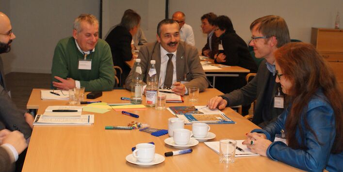 Teilnehmende sitzen am Gruppenarbeitstisch und sprechen miteinander. Auf dem Tisch liegen Moderationsmaterialien wie Stifte und farbige Moderationskarten sowie Getränke. Im Hintergrund sieht man andere Teilnehmende ebenfalls am Gruppenarbeitstisch diskutierend. 