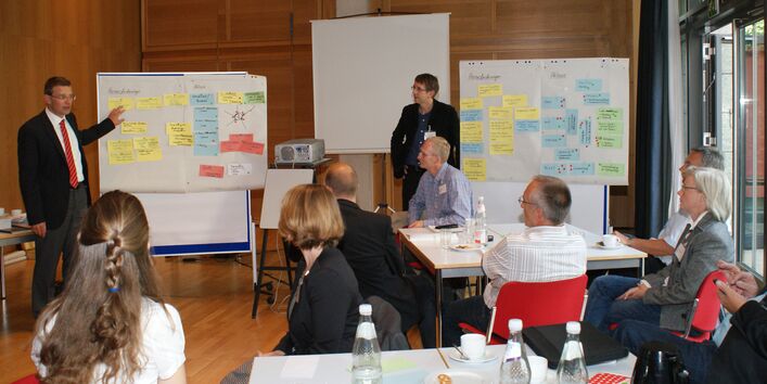 Teilnehmer sitzen an Gruppenarbeitstischen und hören Vortragenden zu. Vortragende stehen im Hintergrund vor Moderationswänden, die mit bunten Moderationskarten bestückt sind. Von der rechten Seite her scheint Tageslicht durch die Fensterfront.