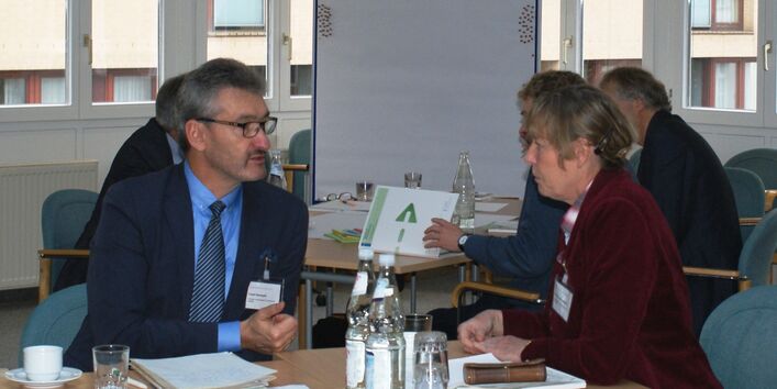 Zwei Teilnehmer befinden sich im Dialog miteinander sitzend am Gruppenarbeitstisch. Im Hintergrund stehen weitere Gruppenarbeitstische vor einer hellen Fensterfront.