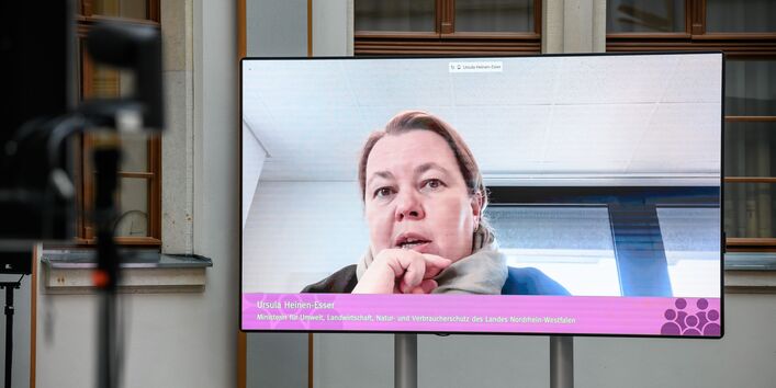 In dem hellen historischen Innenhof zeigt sich eine Teilnehmende auf dem großen flachen Bildschirm sprechend und nachdenklich. Sie hält sich die rechte Hand mit leicht gebeugten Fingern vor das Kinn.