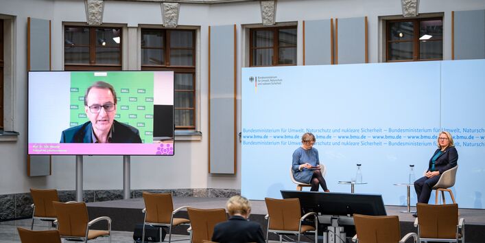 Der Blick geht in dem hellen historischen Innenhof von hinten über das Publikum hin zum Podium vor blauem Hintergrund, wo die Umweltministerin und eine Konferenzteilnehmerin sitzen im Gespräch mit dem Präsidenten des Umweltbundesamtes, der links des Podiums auf dem großen flachen Bildschirm zugeschaltet wurde.