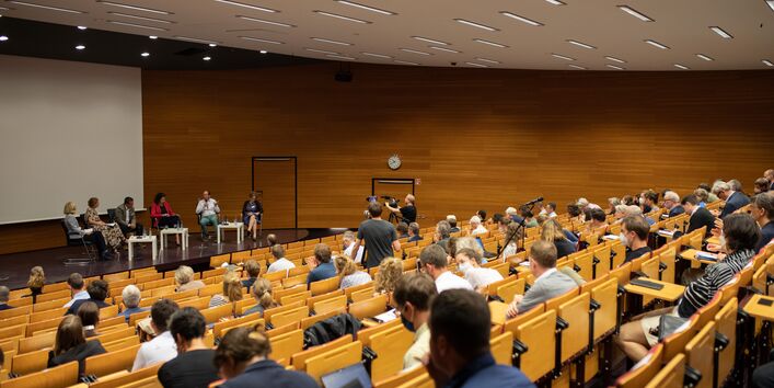Der Blick geht über das Publikum hinweg zu der Bühne, wo die Podiumsteilnehmden vor den niedrigen hellen Tischen sitzen. Ein Teilnehmender spricht in das Handmikrofon.