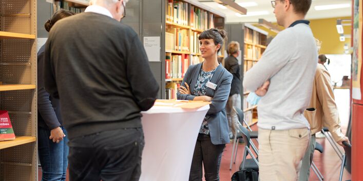 Workshopteilnehmende stehen um einen Stehtisch und tauschen sich aus. Der Blick geht auf eine Teilnehmerin mit verschränkten Armen.