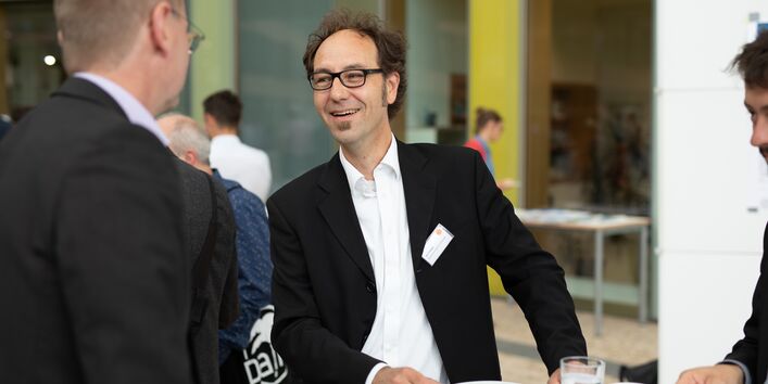 Teilnehmende in dunkler Kleidung unterhalten sich im Atrium am Veranstaltungsort vor dem Hintergrund einer mit Holz durchsetzten und verglasten Front.