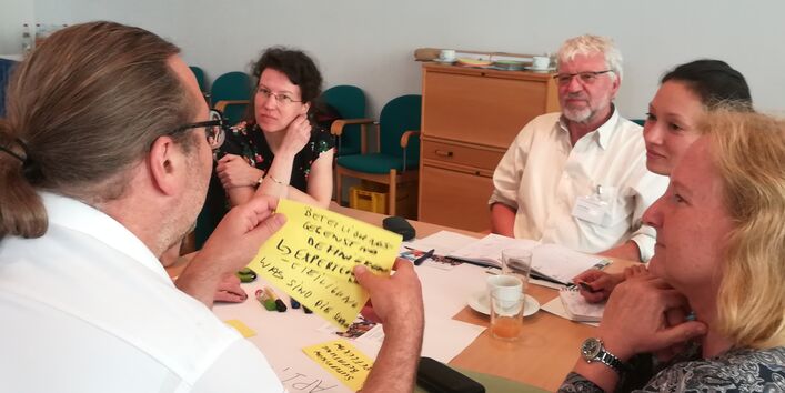 Teilnehmende sitzen am Gruppenarbeitstisch und hören einem Teilnehmenden zu. Er liest Arbeitsergebnisse von gelben Moderationskarten vor, die er in den Händen hält. Wir schauen ihm als Bildbetrachtende über die Schulter.