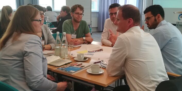 Teilnehmende sitzen im angeregten Gespräch miteinander am Gruppenarbeitstisch. Auf dem Tisch liegen Materialien wie Papier, Stifte, bunte Blätter. Es stehen Getränkeflaschen mit Wasser auf den Tischen sowie Tassen und Gläser.