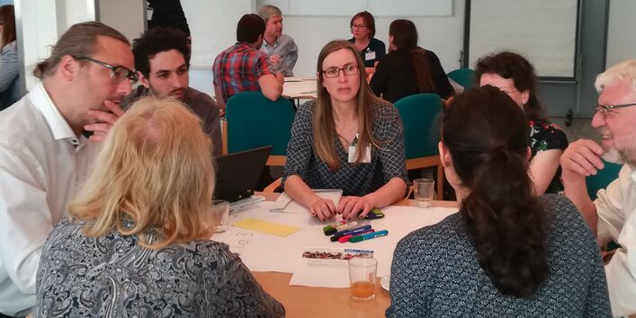 Teilnehmender unterhalten sich angeregt miteinander. Sie sitzen dicht beieinander an einem Gruppenarbeitstisch.