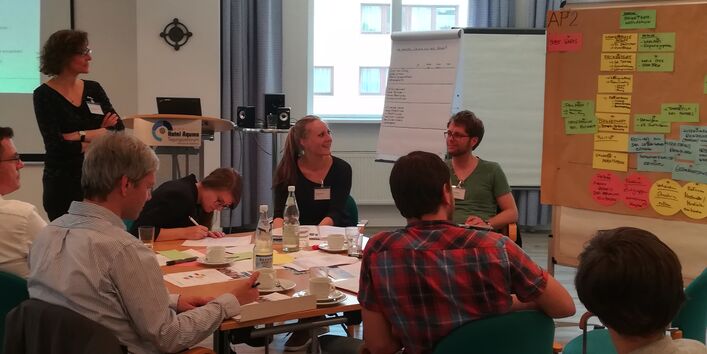 Teilnehmende sitzen in einer großen Runde am Gruppenarbeitstisch vor der Moderationswand mit den Arbeitsergebnisse auf farbigen Moderationskarten. Sie diskutieren angeregt und lachen miteinander.